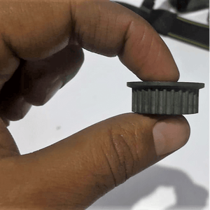 3mm Timing Pulley P28 - a close-up shot of a small, grey, cylindrical toothed pulley held between two fingers.