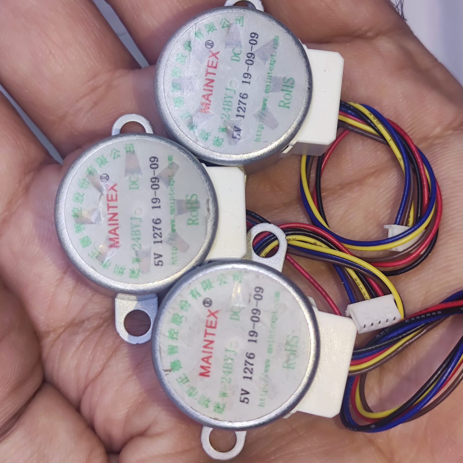 Arduino 5V Stepper Motor 24BYJ - three silver circular motors with white plastic connectors and multi-coloured wiring held in a hand.
