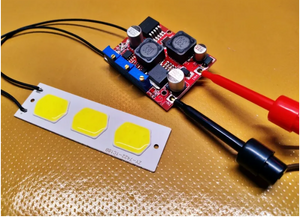 DC-DC Boost & Buck Voltage Converter LM2577 & LM2596 Module - & Buck Voltage Converter LM2577 & LM2596 Module connected to a three-LED COB strip with red and black test probes attached to the input.