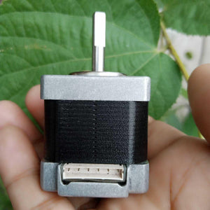 NEMA 11 stepper motor without connector - a silver metal body, a flat-sided shaft, and a white six-pin connector port, held in a hand against a green leaf background.