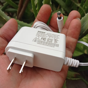 New High Quality 220V To 12V 1A AC DC Adapter - a white wall-plug power supply with a barrel connector, held in a hand against a green plant background.