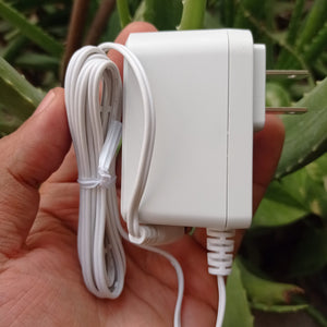 New High Quality 220V To 12V 1A AC DC Adapter - a white wall-plug power supply with a long white cable held in a hand against a blurred plant background.