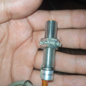 Omron Inductive Proximity Sensor Switch E2e-X2E1 - a cylindrical silver metal sensor with a threaded body, two nuts, and a washer held in a hand.