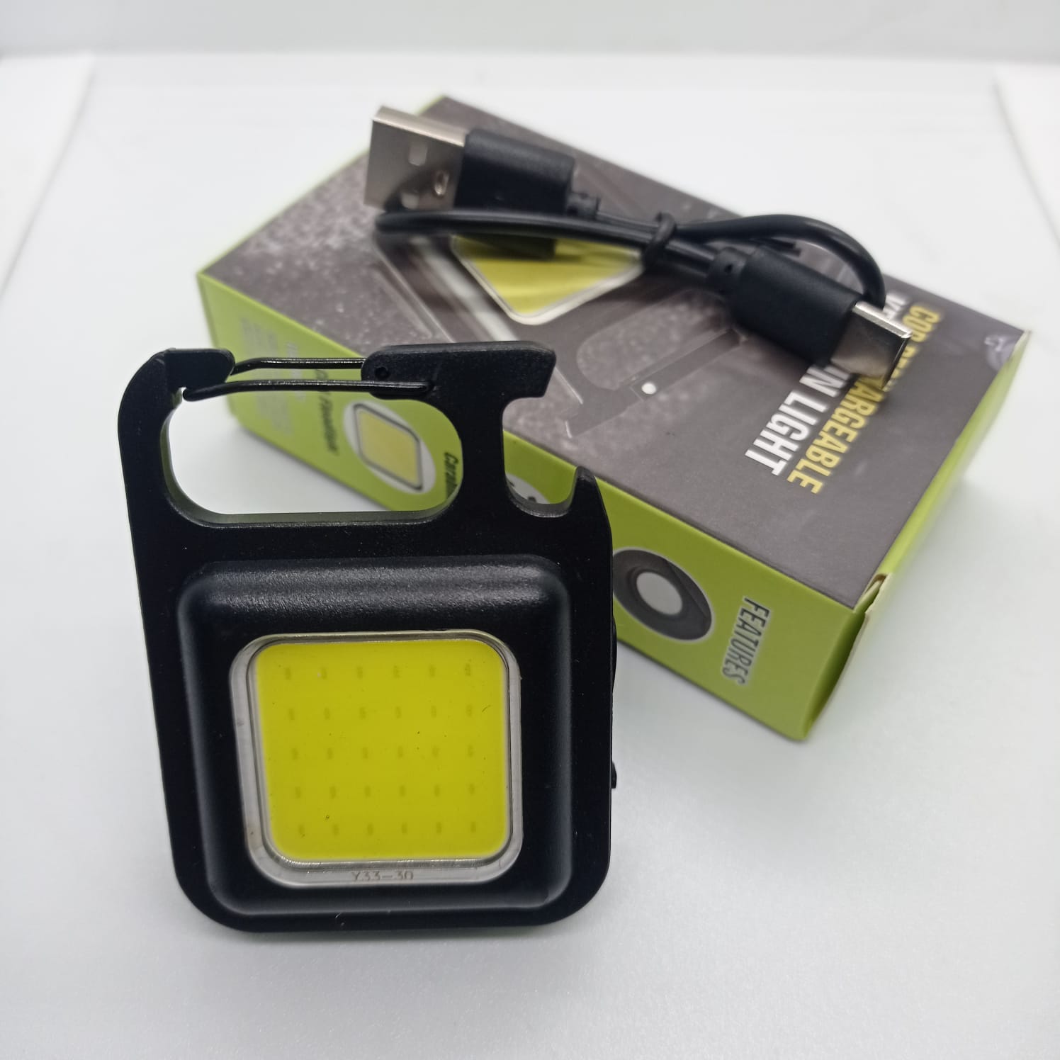 Portable COB Flashlight - Emergency Light with Rechargeable Keychain - Durable Plastic Casing - black square casing, integrated carabiner, and a USB charging cable resting on its product packaging box.