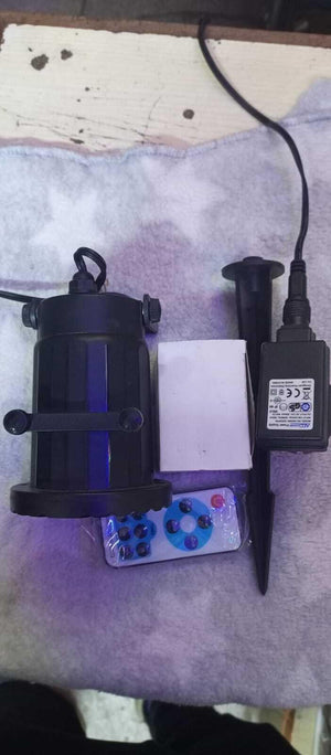 Projector - shown with a black cylindrical light housing, a power adapter with a ground stake, a small remote control, and a white box.