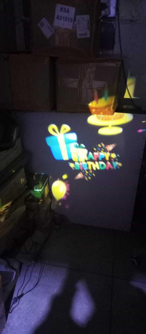 Projector - Projector projecting a colorful "Happy Birthday" graphic with a cake, gift box, and balloon onto a wall below cardboard boxes.