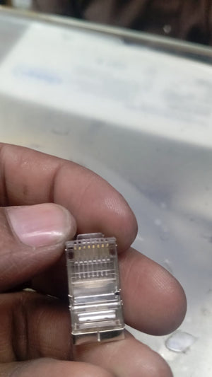 RJ45 Connector with meteal casing - with metal casing, shown in a close-up view held by fingers, displaying the transparent plastic housing and the eight internal gold pins.