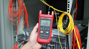 UNI-T UT692G High-Power Optical Power Meter – -50~+26DBM, 800–1700nm, FC/SC/ST, InGaAs, ±5%, IP65, Fiber Optic Power Meter - UNI-T UT692D optical power meter held in a hand, showing a red handheld device with a digital screen reading -70.00 dBm and connected to yellow fiber.