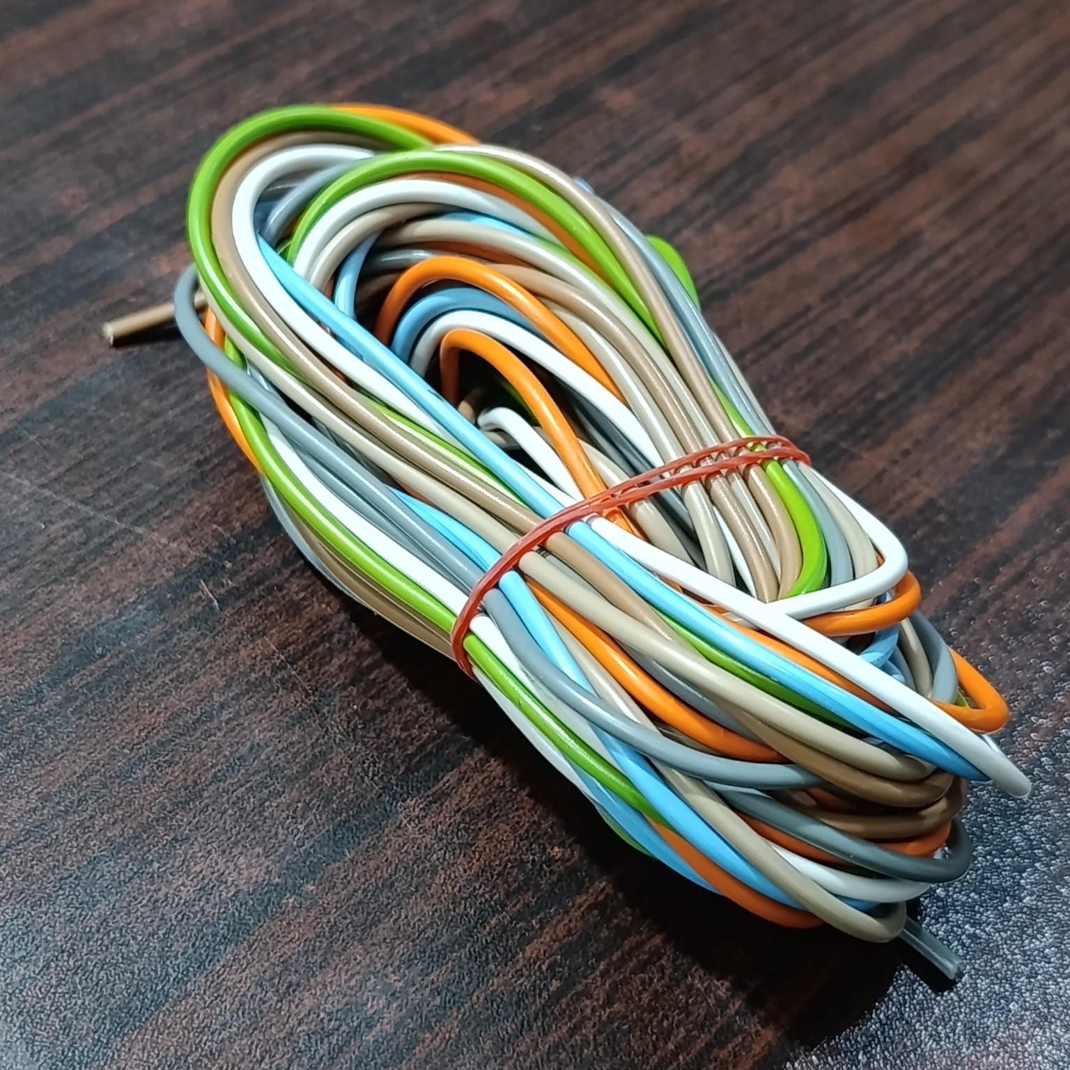 1 Meter 7 Core Wiring Cable - shown as a coiled bundle of multi-coloured insulated wires held together by a thin red rubber band on a wooden surface.