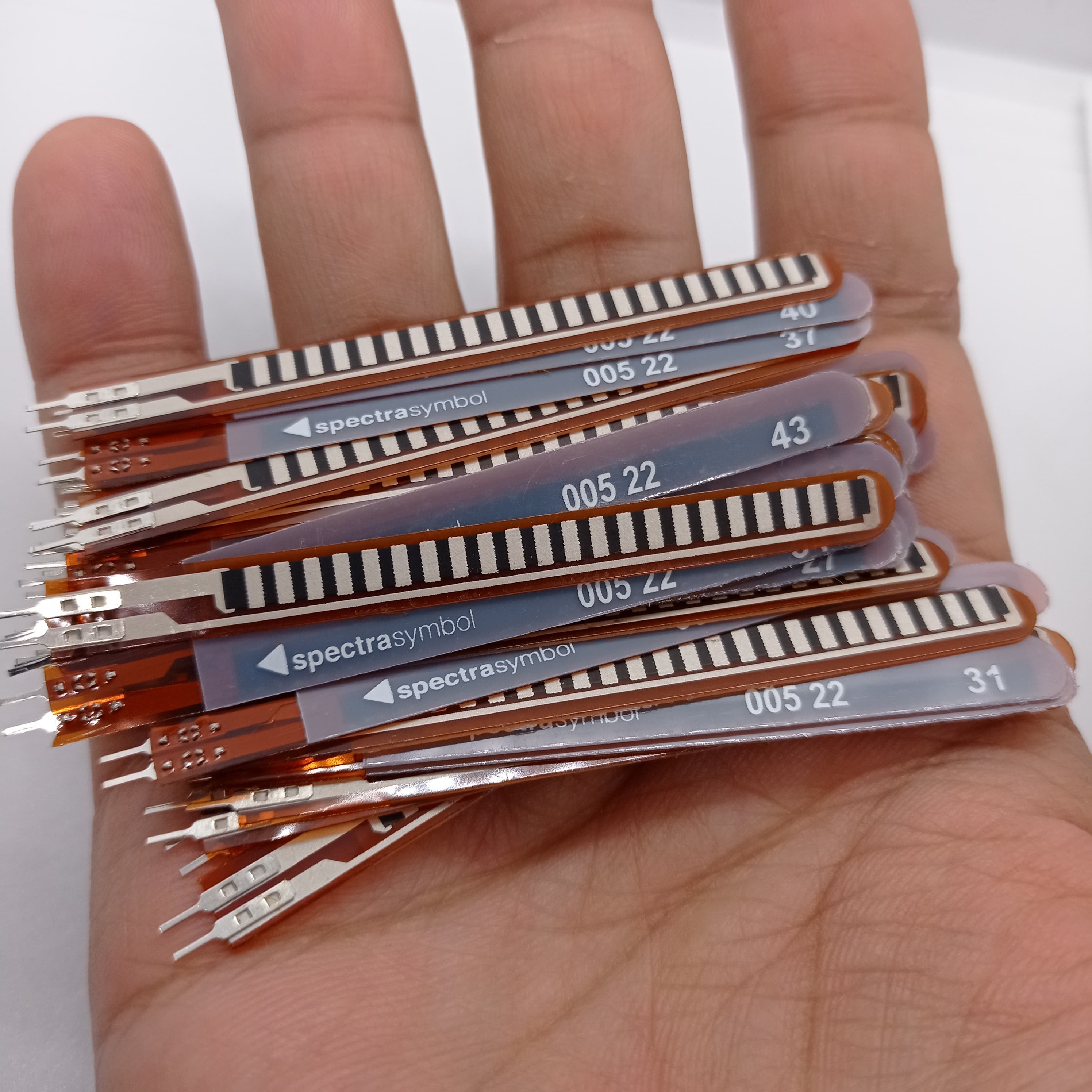 1 Piece 2.2 Inch Flex Sensor Bend Detection Sensor - a hand holding a stack of thin, brown, flexible strip sensors with metal pin connectors.