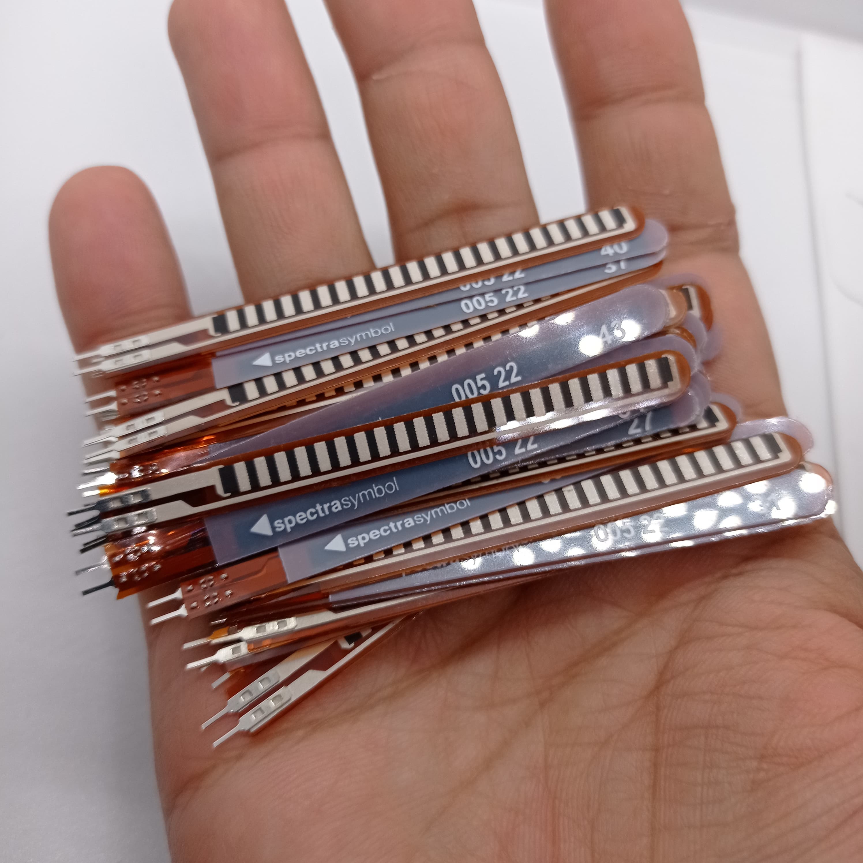 1 Piece 2.2 Inch Flex Sensor Bend Detection Sensor - a hand holding a stack of thin, brown, flexible strip sensors with metallic connectors and printed "spectrasymbol" labels.