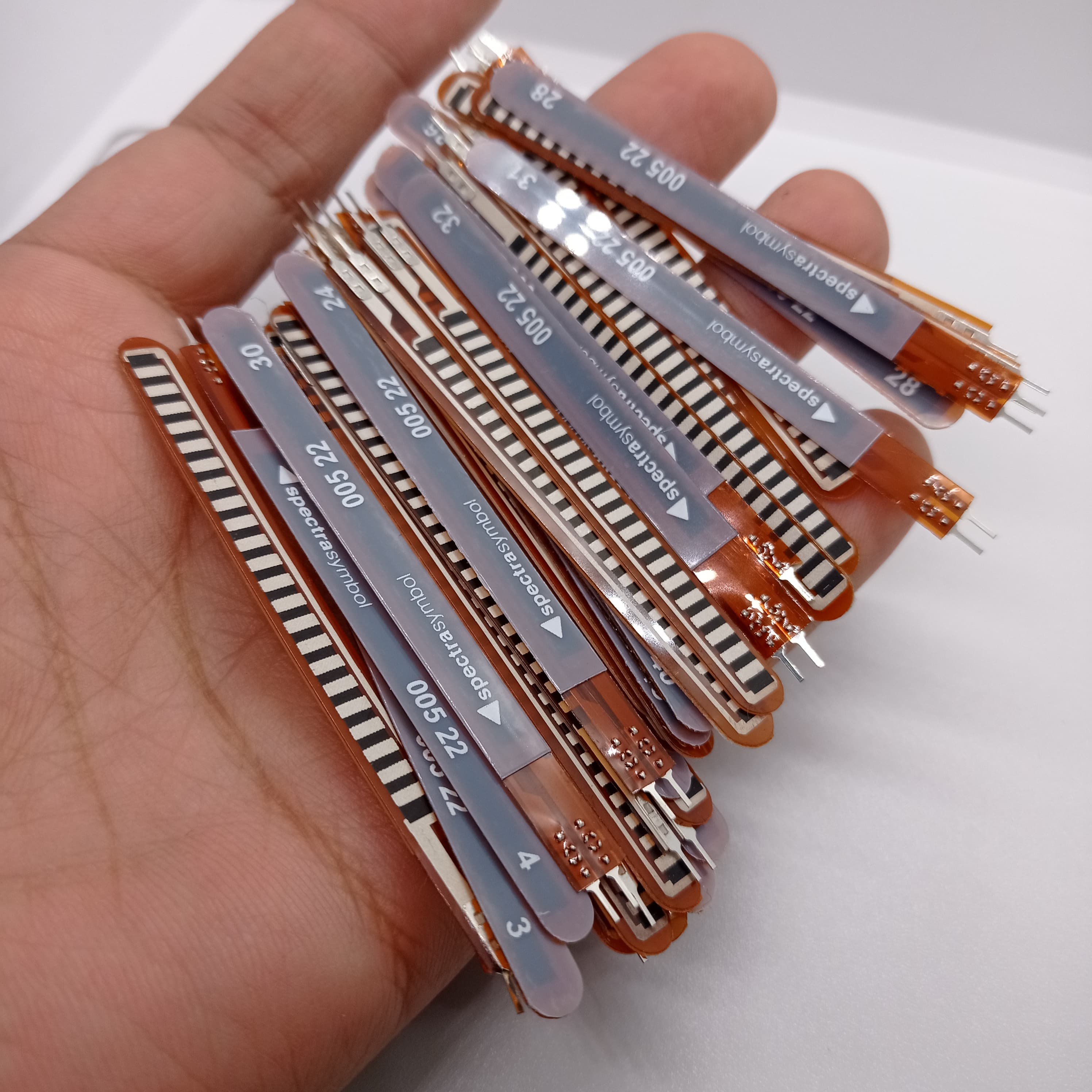 1 Piece 2.2 Inch Flex Sensor Bend Detection Sensor - a hand holding a stack of multiple long, thin, flexible brown and grey sensors with printed labels and connector pins.