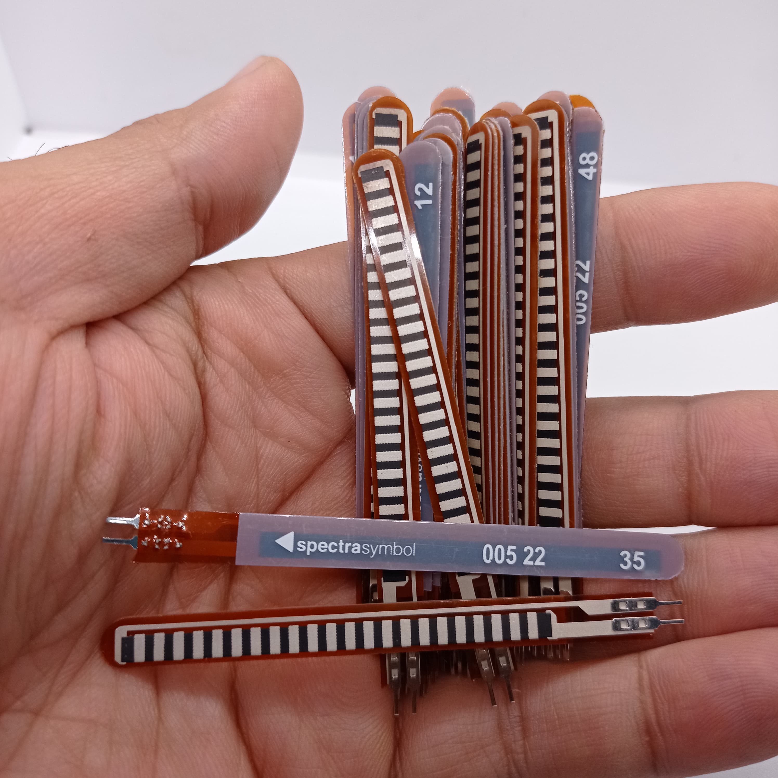 1 Piece 2.2 Inch Flex Sensor Bend Detection Sensor - a hand holding a bundle of thin, brown, strip-shaped sensors with white connector pins.