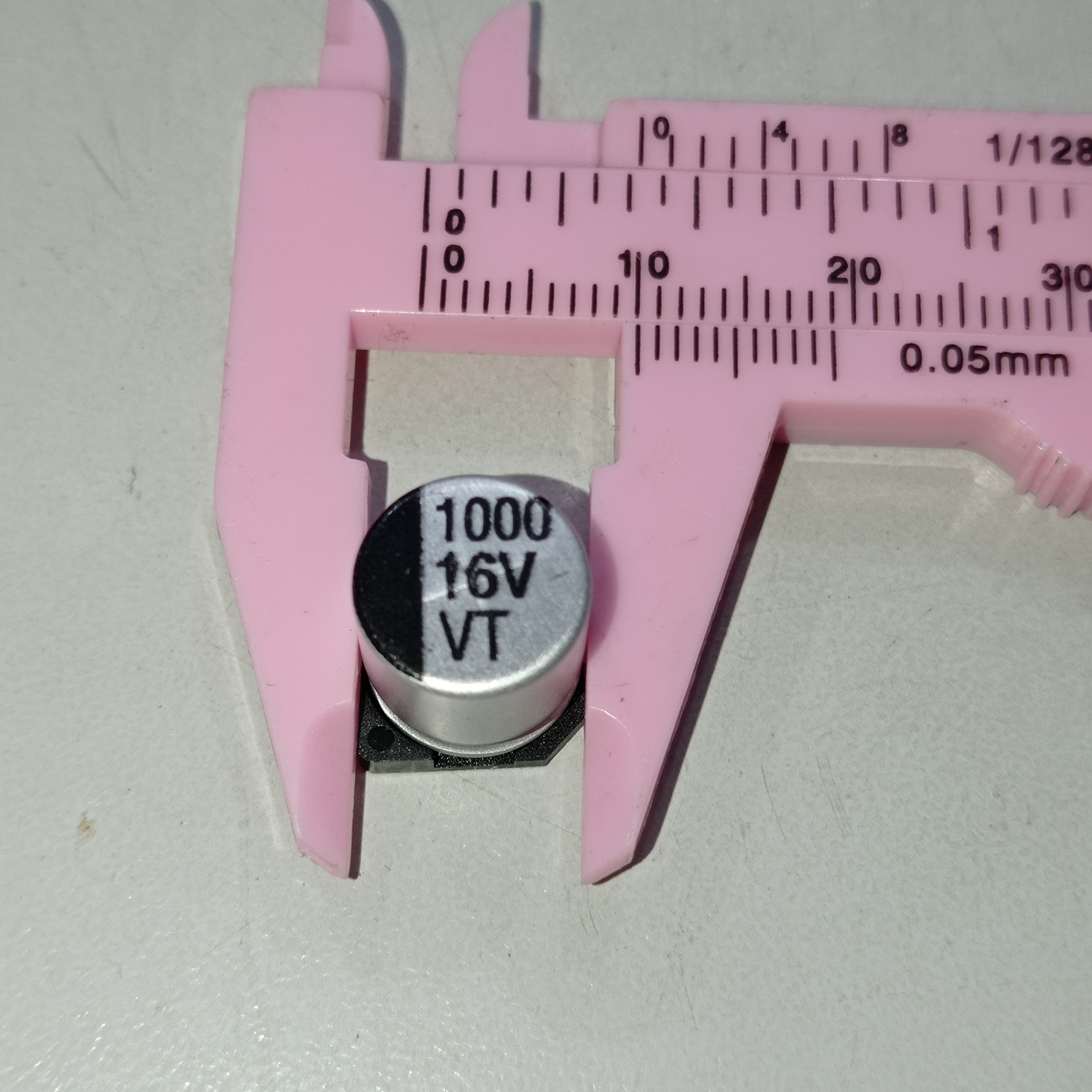 1000uF 16V SMD Capacitor D10XL10.2mm Aluminum Electrolytic Capacitors - SMD - shown in a close-up view being measured by a pink plastic vernier caliper.