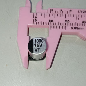 1000uF 16V SMD Capacitor D10XL10.2mm Aluminum Electrolytic Capacitors - SMD - shown in a close-up view being measured by a pink plastic vernier caliper.