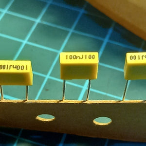 100NJ100 0.1uF 100V Radial Polyester Capacitor - a close-up of three yellow rectangular capacitors with radial leads attached to a paper strip.