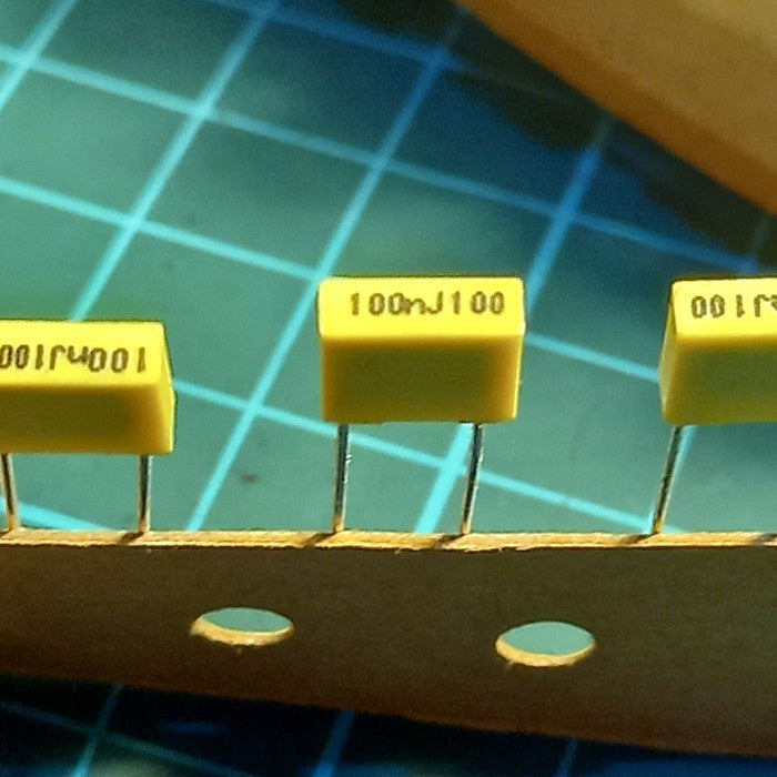 100NJ100 0.1uF 100V Radial Polyester Capacitor - a close-up of three yellow rectangular capacitors with radial leads attached to a paper strip.