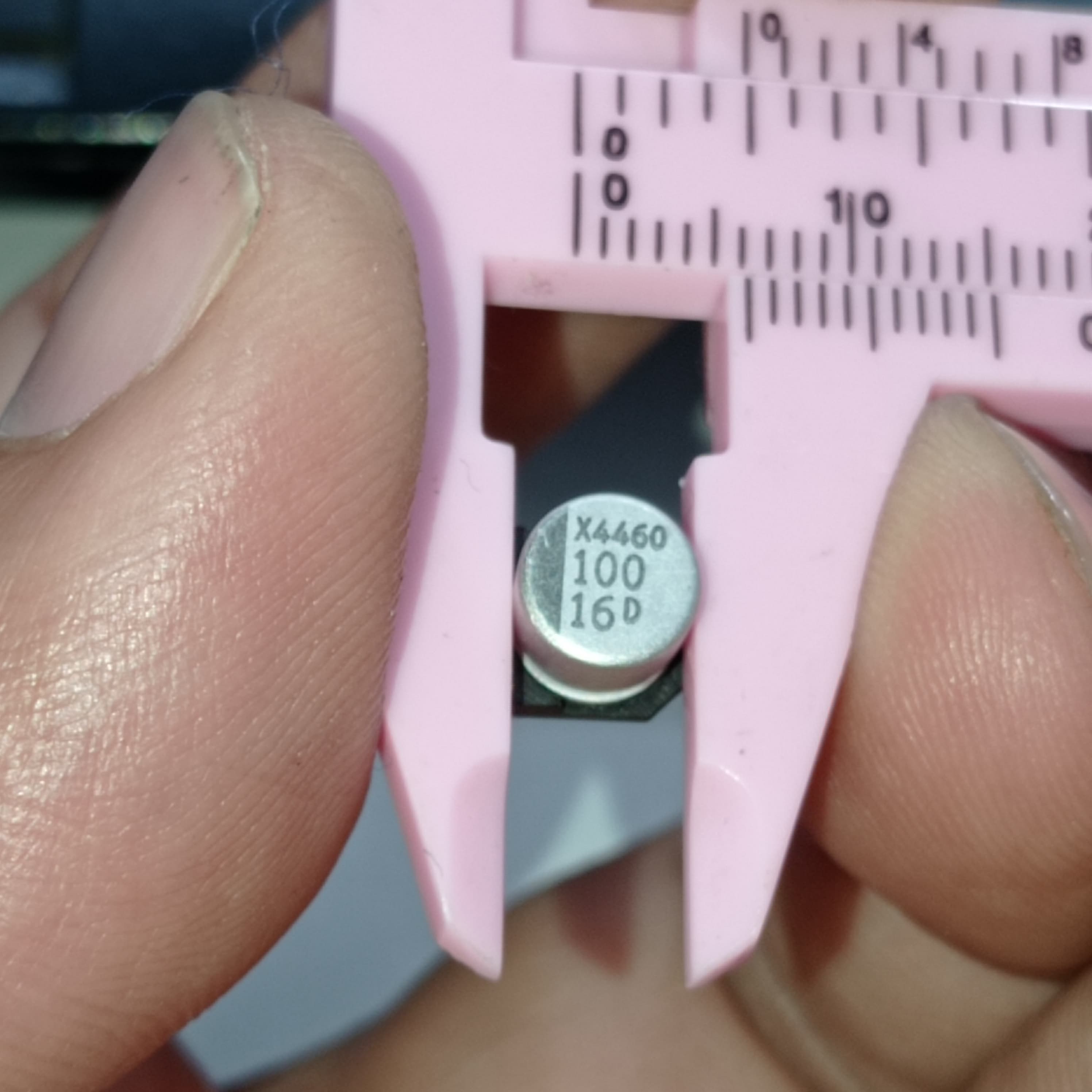 100uF 16V ±20% SMD D6.3XL5.4mm Aluminum Electrolytic Capacitor - shown in a close-up being measured by pink plastic calipers, with "X4460 100 16D" printed on its top.