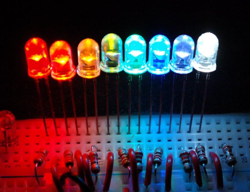 10mm Green LED Light Emitting Diode - shown in a row of eight multi-coloured LEDs glowing on a breadboard with connected resistors.
