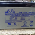12V 9A DC Battery - shown as a rectangular, grey Leoch DJW12-9.0 lead-acid battery with black top terminals and printed technical specifications.