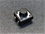 12x12x9mm Tactile Push Button Switch - a square black electronic component with a central circular button and four metal pins on a grey textured surface.