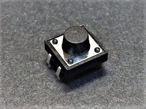 12x12x9mm Tactile Push Button Switch - a square black electronic component with a central circular button and four metal pins on a grey textured surface.