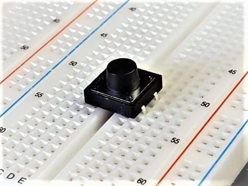 12x12x9mm Tactile Push Button Switch - a black square-shaped tactile button with a round top, mounted on a white breadboard.