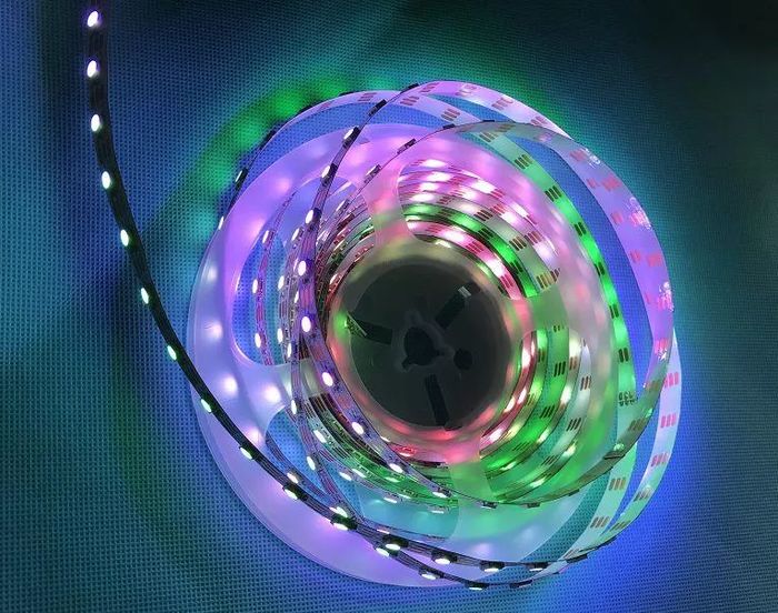 2812 LED Strip - coiled on a plastic spool, glowing with multi-coloured light against a blue textured background.