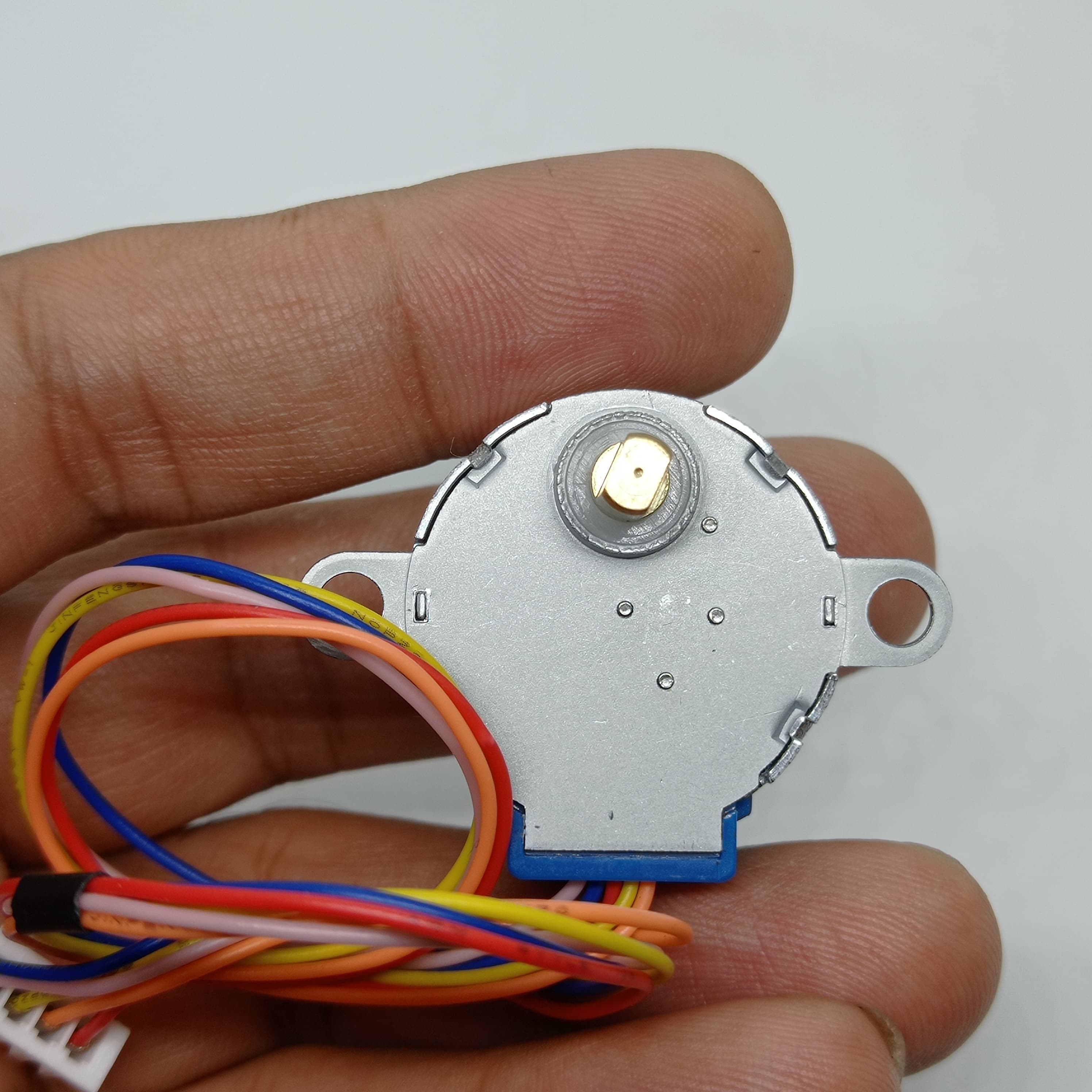 28BYJ-48 5V Stepper Motor - The Best Stepper Motor For Your Projects - held in a hand, showing the silver metal casing, a small brass gear, and a multi-coloured wire harness with a white connector.