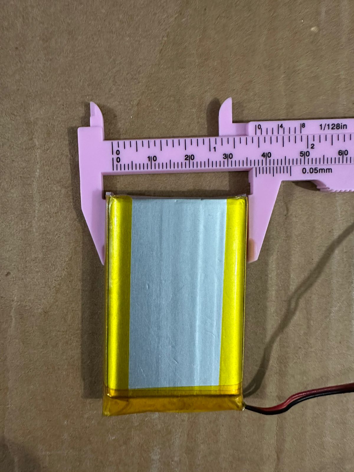 3.7V 2500mAh Lithium-Ion Rechargeable Battery - Li-Po Battery Pack - shown being measured with a pink plastic vernier caliper on a brown cardboard surface.