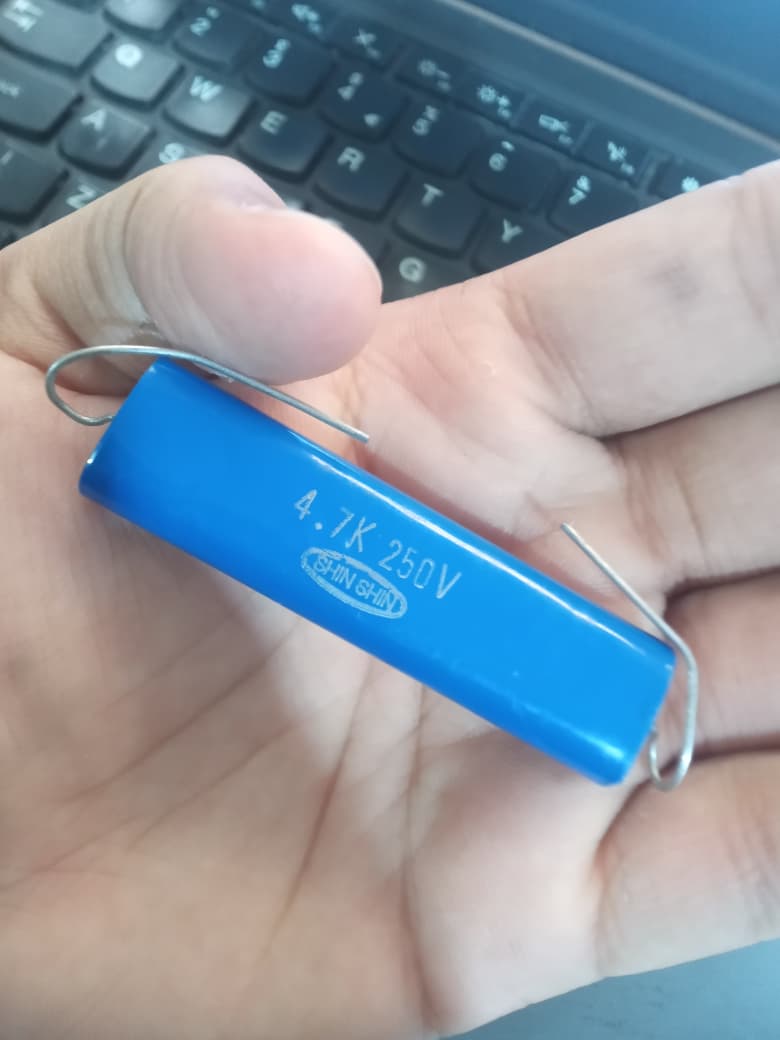 4.7k 250V capacitor - a blue cylindrical electronic component with two wire leads, held in a hand above a computer keyboard.