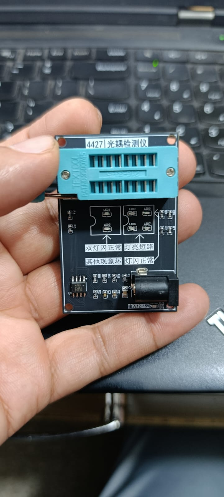 44271 Optocoupler Tester - Photocoupler Testing Module With ZIF Socket - a black circuit board with a blue ZIF socket, a DC power jack, and text labels in Chinese, held in a person's hand.