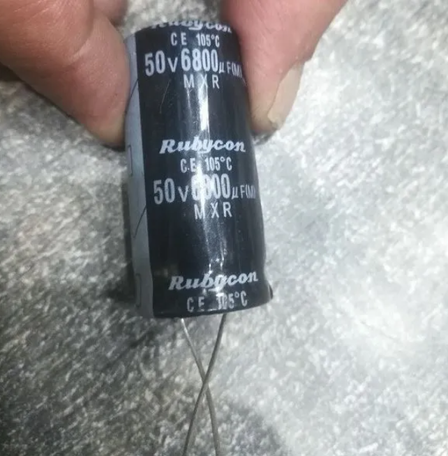 6800uF 50V Electrolytic Capacitor - held by fingers, showing a black cylindrical Rubycon electrolytic capacitor with white printed text and two wire leads.