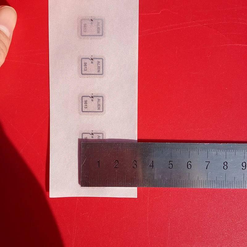 915MHz Uhf Mini RFID Tag - shown in a strip of four white square labels with black text, measured against a metal ruler on a red background.