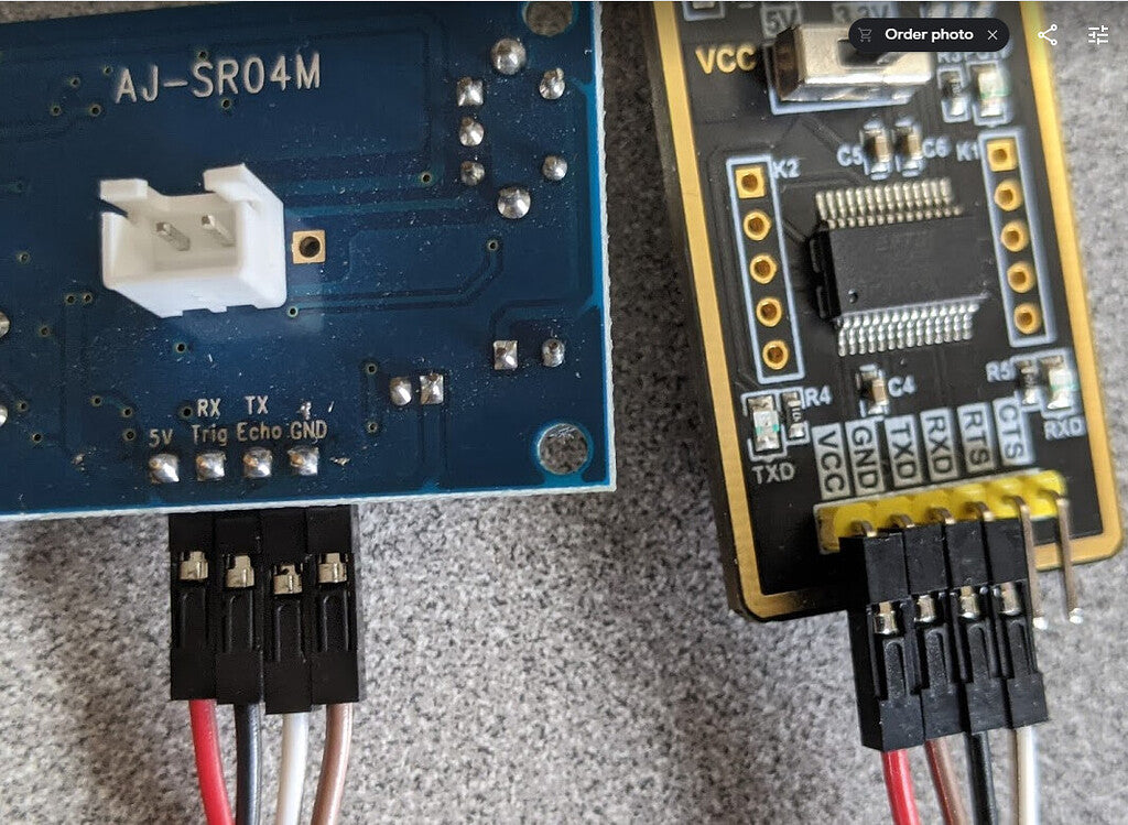 AJ-SR04M V3.0 Waterproof Ultrasonic Distance Sensor - connected by jumper wires to a black USB-to-TTL serial adapter board on a grey surface.