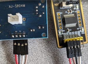 AJ-SR04M V3.0 Waterproof Ultrasonic Distance Sensor - connected by jumper wires to a black USB-to-TTL serial adapter board on a grey surface.
