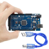 Arduino Mega 2560 R3 - held in a hand, featuring a blue circuit board with ports and buttons, accompanied by a blue USB cable.