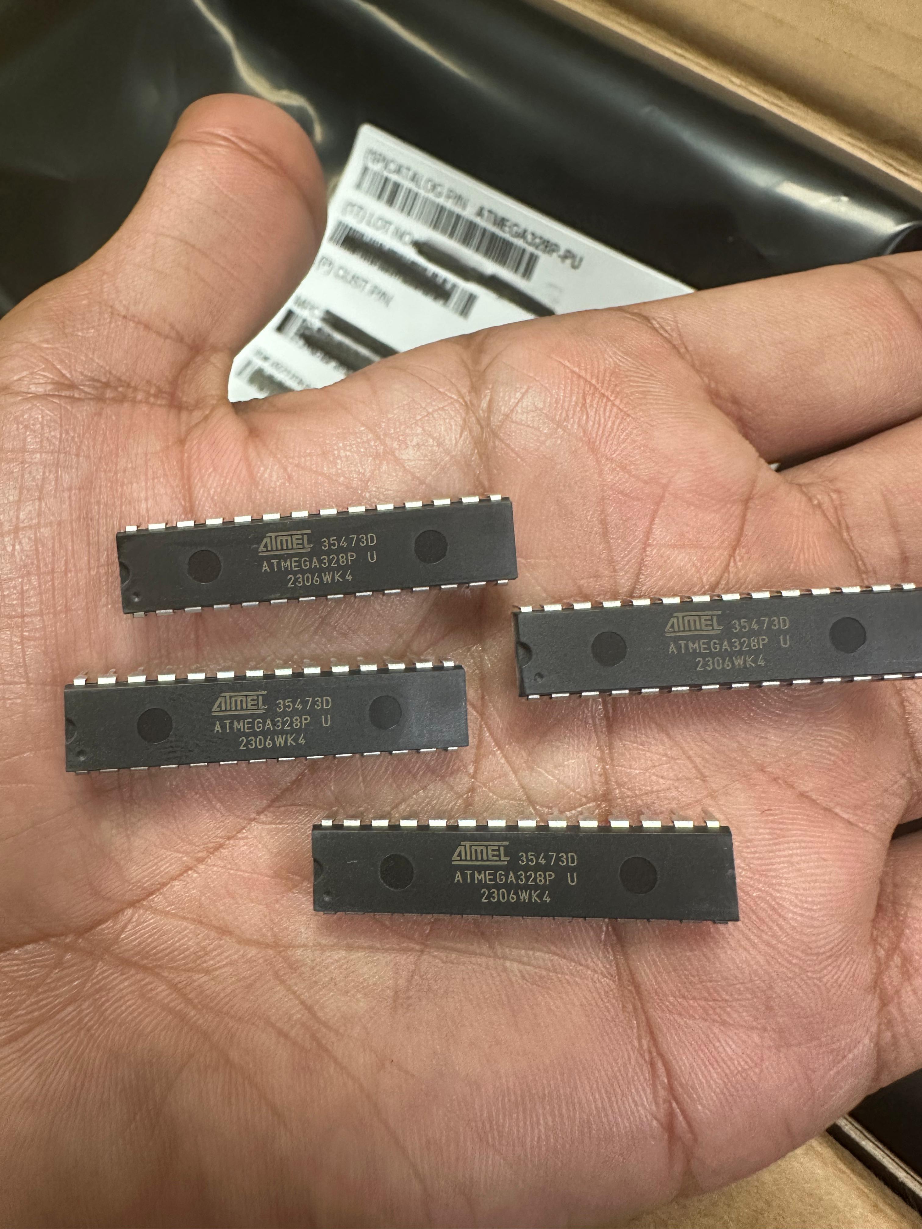 ATMEGA328P-U Microcontroller | High-performance MCU | Digilog.pk - a close-up shot of four black, rectangular 28-pin integrated circuit chips held in a hand, with packaging labels in the background.