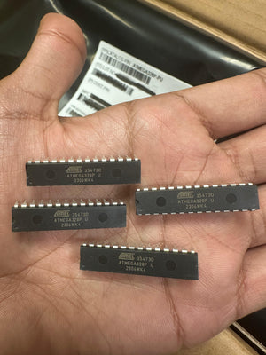 ATMEGA328P-U Microcontroller | High-performance MCU | Digilog.pk - four black 28-pin DIP integrated circuit chips held in a hand with a blurred product label in the background.