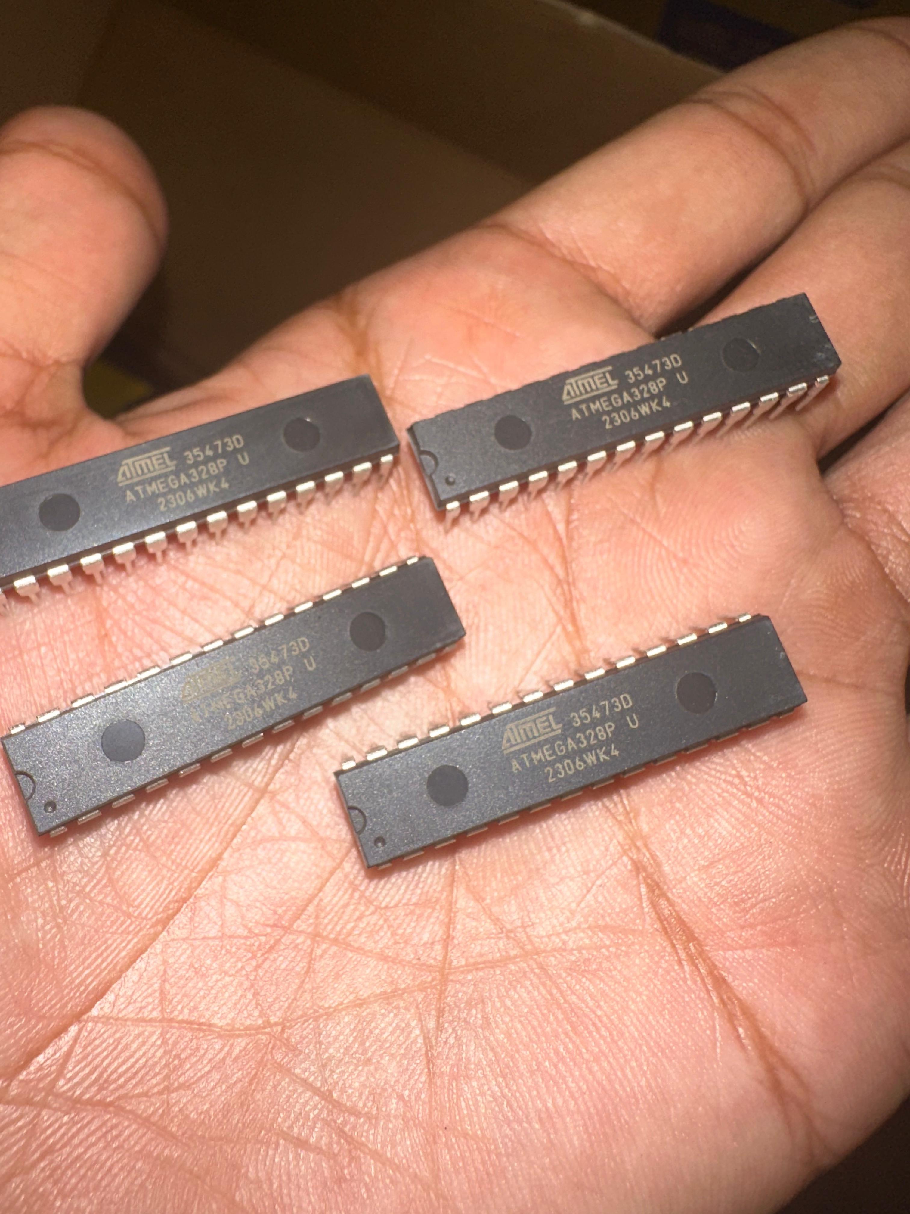 ATMEGA328P-U Microcontroller | High-performance MCU | Digilog.pk - four black rectangular 28-pin DIP integrated circuits held in a hand, each marked with Atmel and ATMEGA328P U 2306WK4.