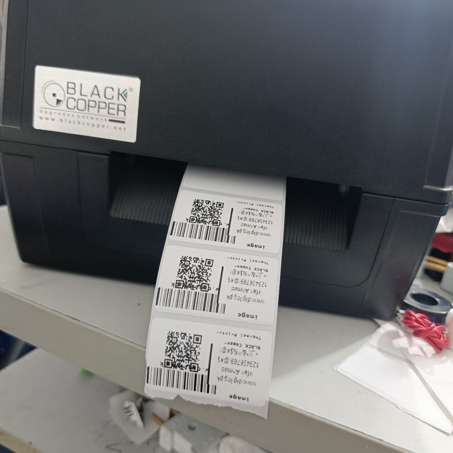 Black Copper Label Designer Software – Offline Package - – Offline Package: a black thermal label printer printing a continuous strip of white labels with QR codes and text.
