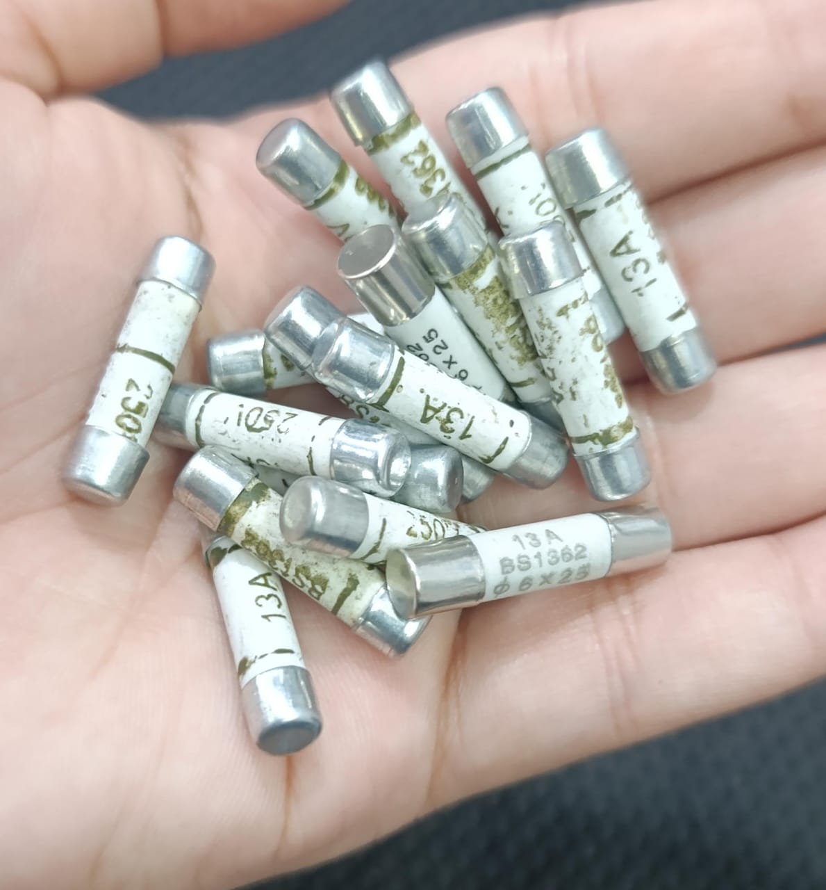 BS1362 13A Ceramic Fuse - shown as a pile of multiple white cylindrical ceramic fuses with metal end caps held in a person's hand.