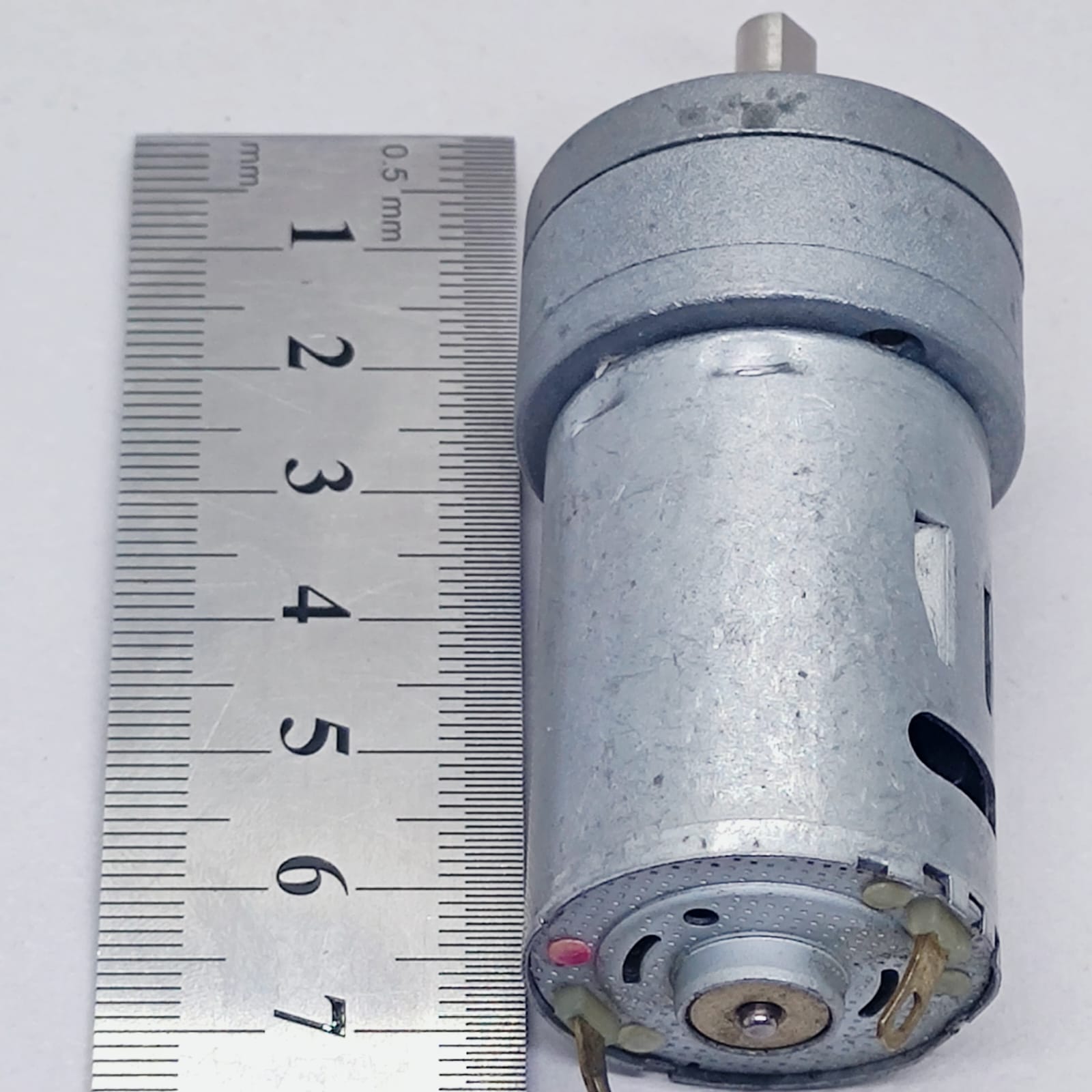 DC Motor DC Electric Geared Motor - shown next to a metal ruler for scale, featuring a cylindrical silver motor body with an attached gear head.