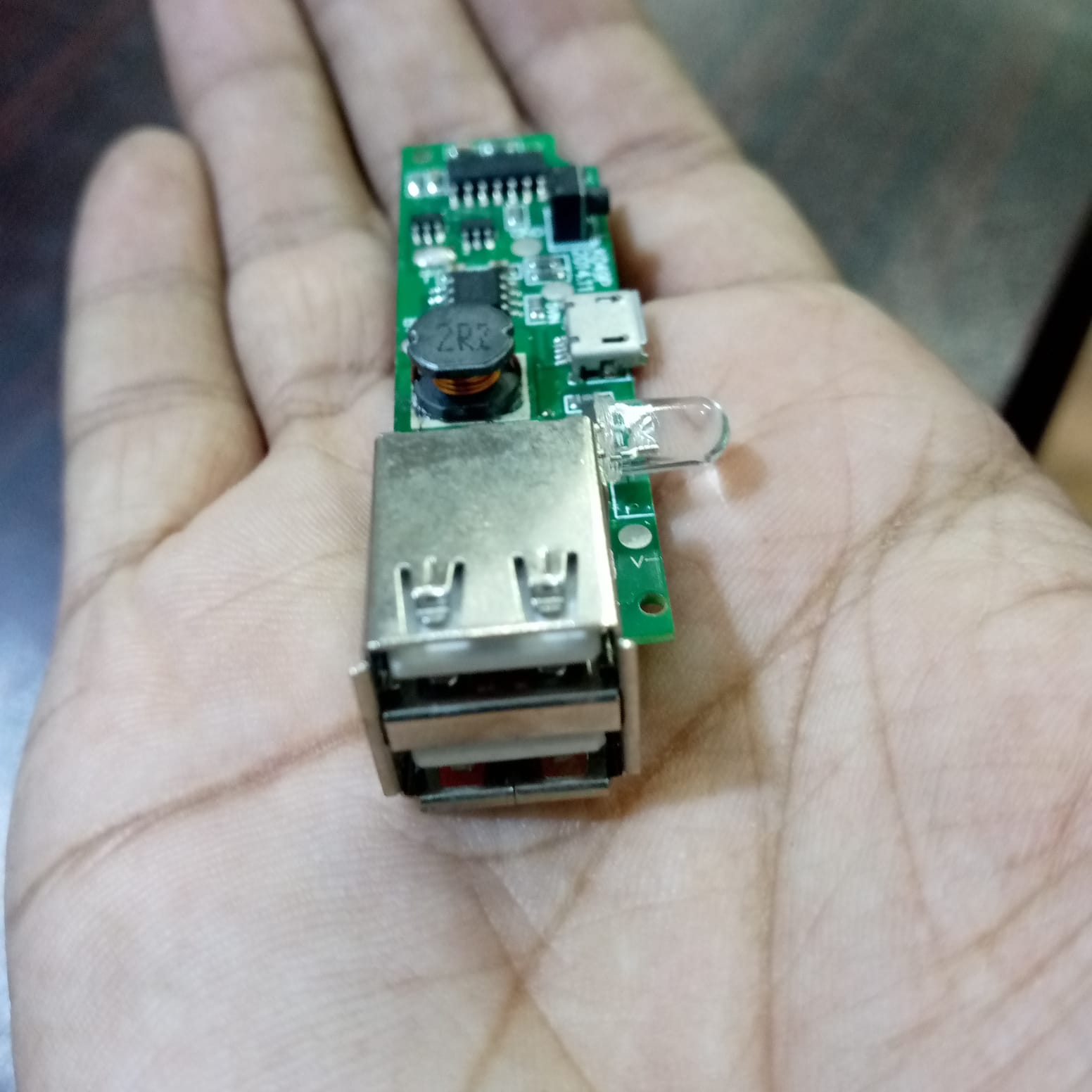 Dual USB Power Bank Module - shown held in a hand, featuring a green circuit board with two USB-A ports, a micro-USB input, and an LED indicator.