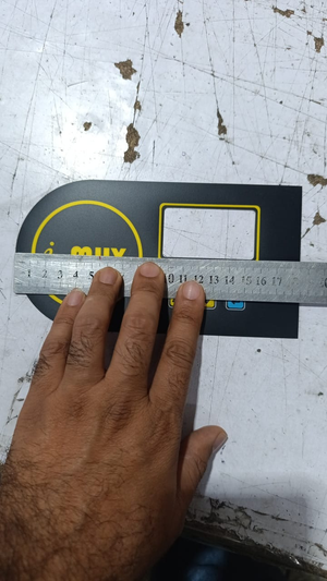 Hybride LCD Sticker Idm-7 Sticker For Graphical LCD Based Local Solar Inverter - a black rectangular panel with a yellow circular logo and a rectangular cutout, being measured with a metal ruler.