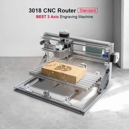 Imported CNC Engraving PCB Milling Machine Wood Carving CNC 3018 - a silver metal 3-axis engraving machine shown carving a Transformers logo into a rectangular wooden block on its platform.