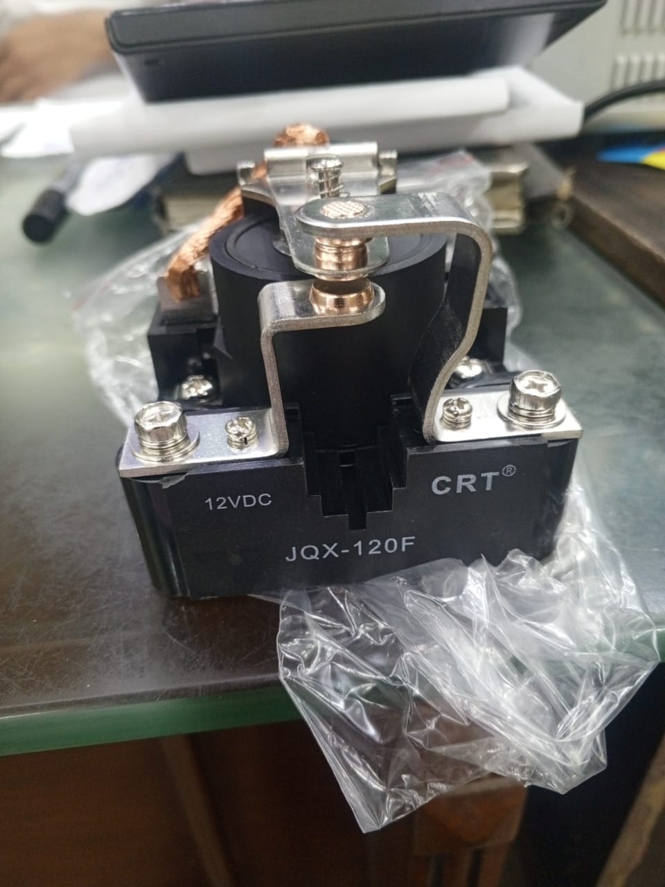 Jqx 120F 12V Relay - a black electromagnetic relay with metal terminals and screws, shown from the front on a desk with plastic packaging.