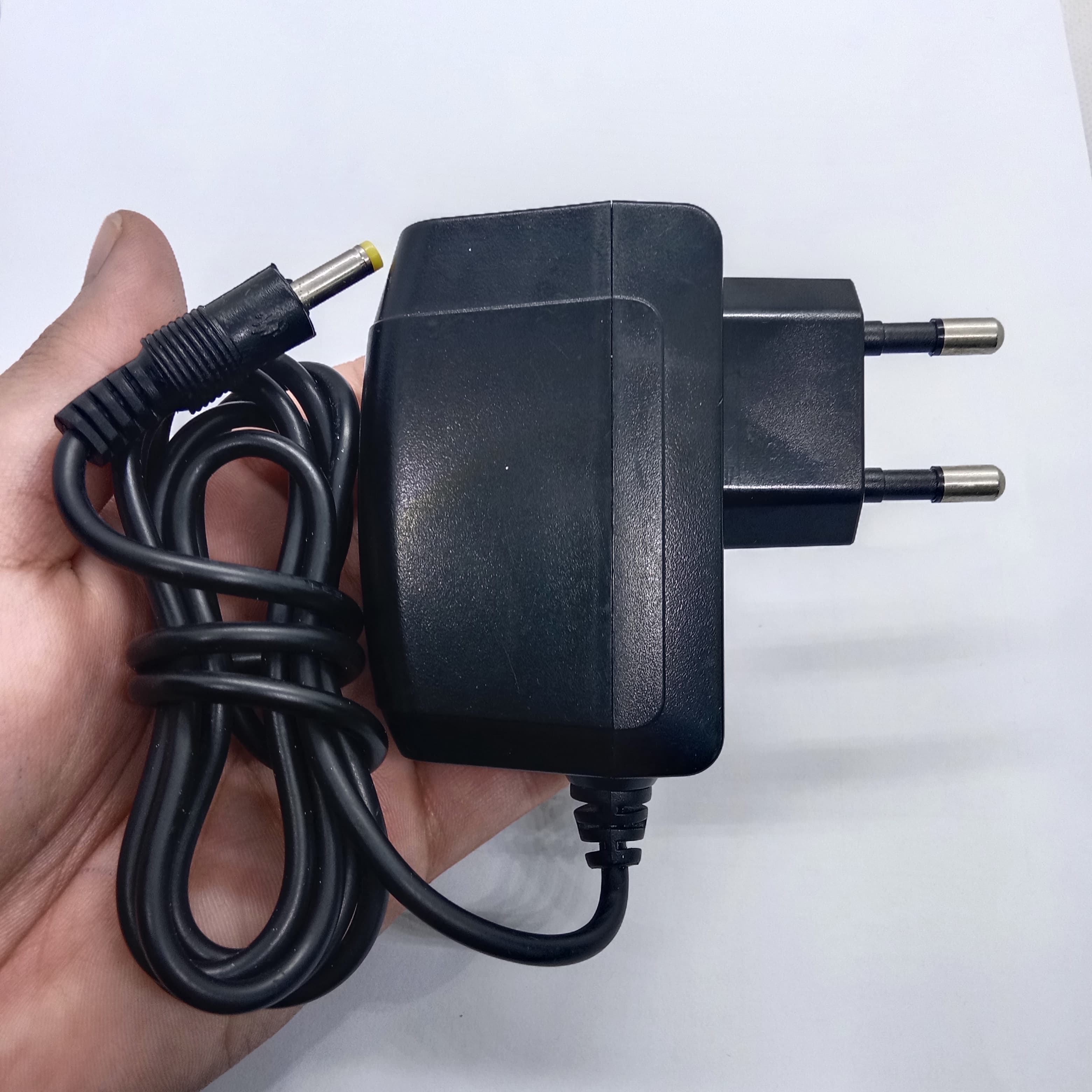 LG U+ 5V 2amp Power Adaptor Kecil Jack - a black wall-plug power supply with a long cable and a yellow-tipped barrel connector held in a hand.