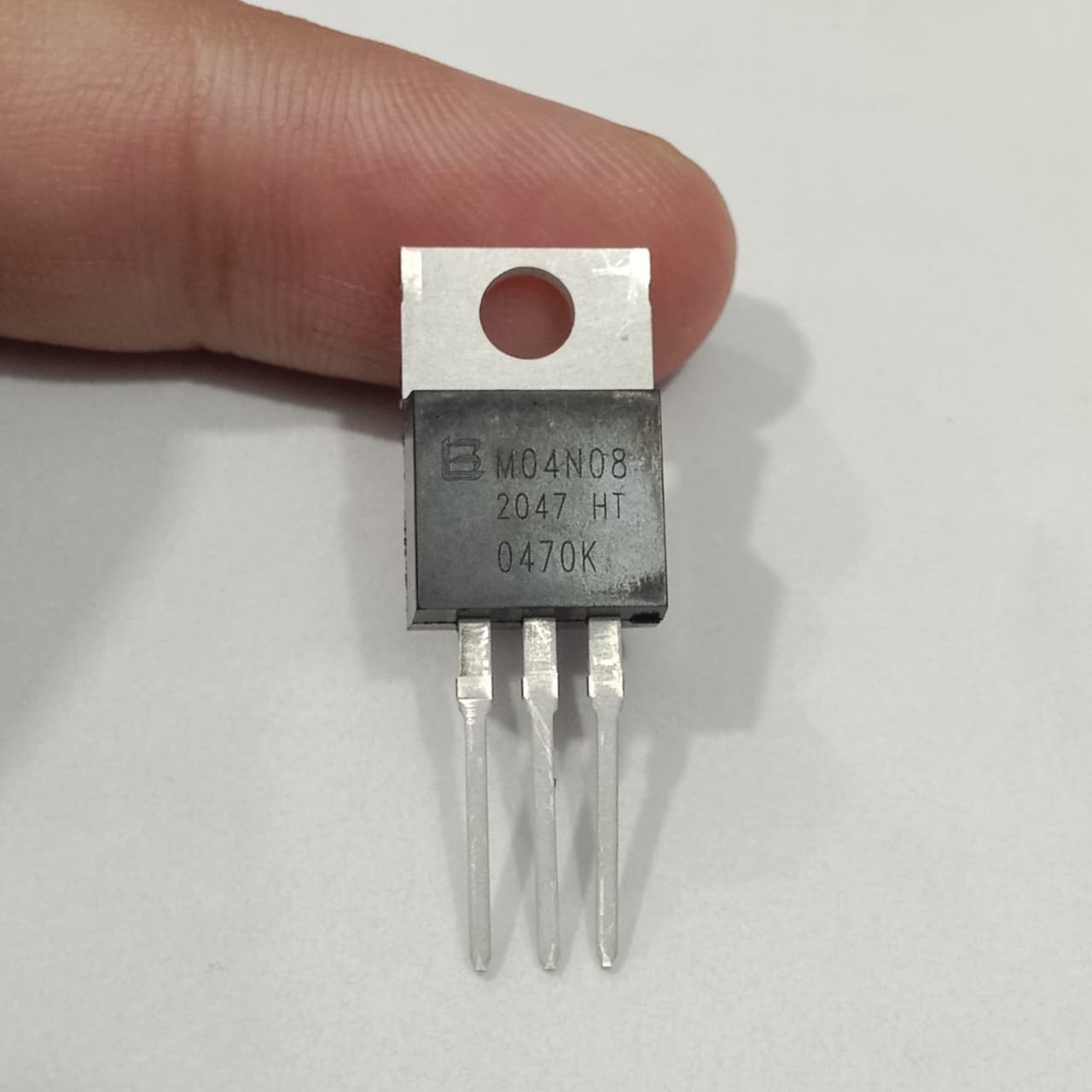 M04N08 N-channel MOSFET - a black, three-pin electronic component with a metal heat sink tab, held by a finger against a plain white background.