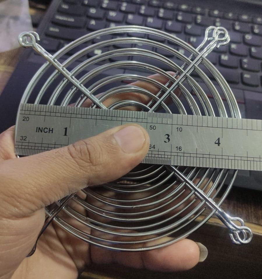 Metal Exhaust Fan Guard Grill – 4 Inch | Protective Jali Cover for Cooling & Ventilation - shown being measured with a metal ruler, displaying its circular silver wire frame and mounting holes.